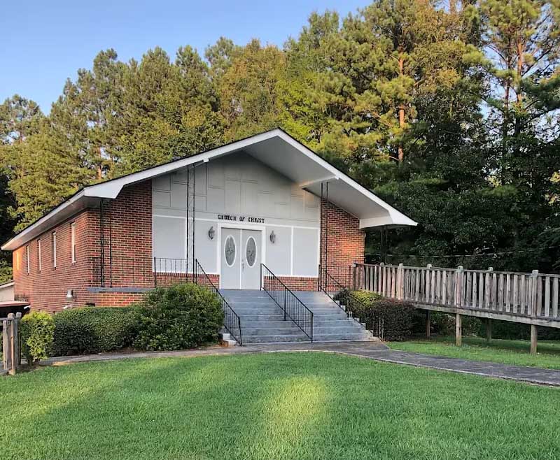 The Saks Church of Christ building: red brick with white double doors, front steps, and a wooden ramp, surrounded by tall pines